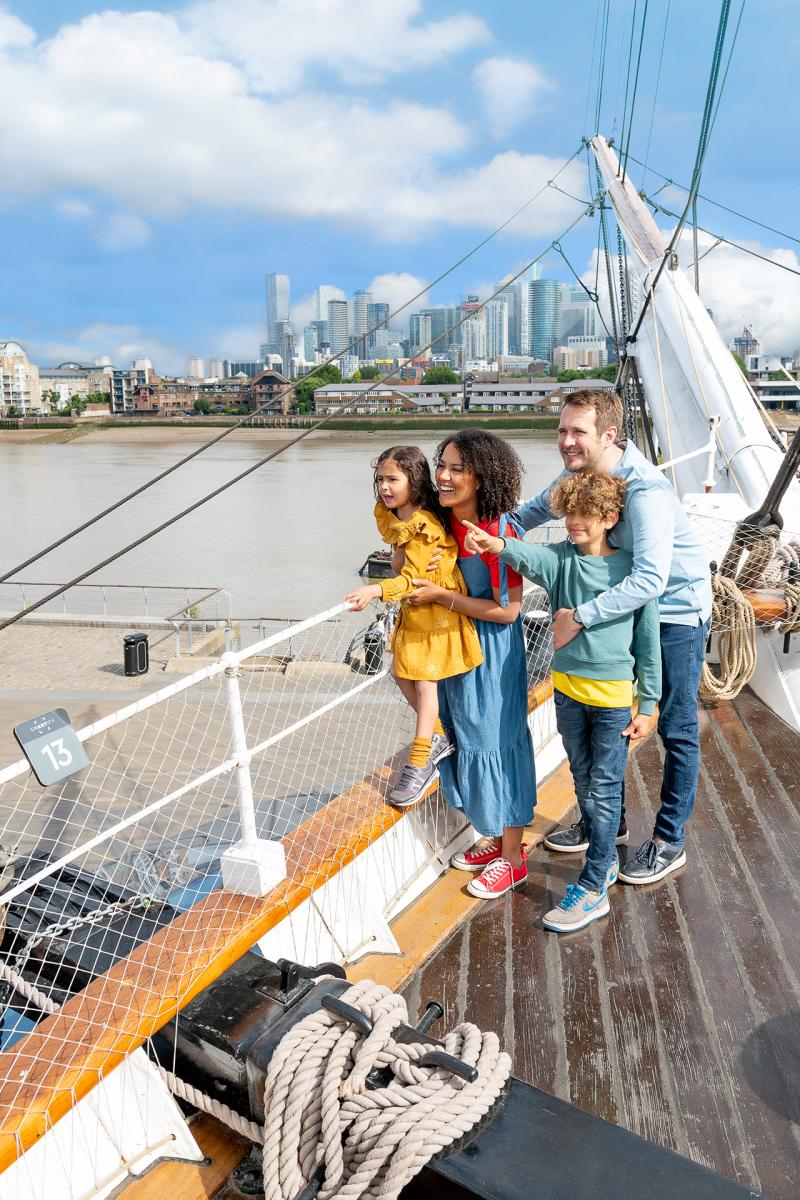 A family stand on the main deck of historic ship Cutty Sark, looking out across Greenwich with the River Thames and skyscrapers of Canary Wharf shining in the background