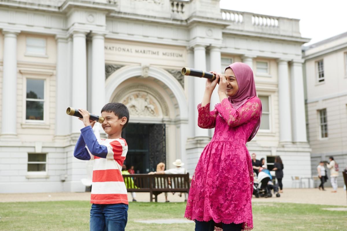Two children look through telescopes outside the National Maritime Museum. They are smiling as they peer through the historic items