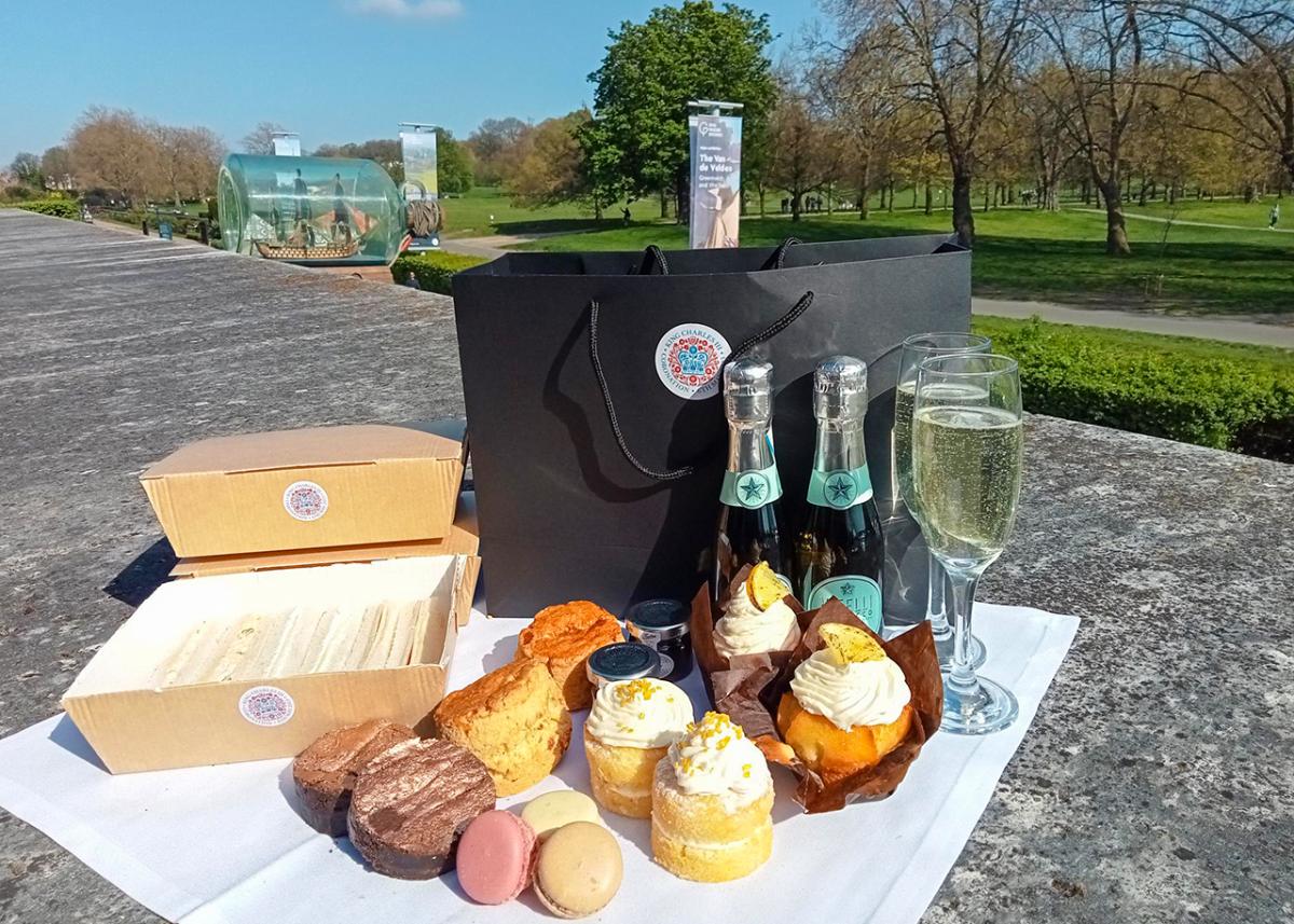 A picnic hamper laid out in front of Greenwich Park, including a box of mini sandwiches, brownies, macaroons, scones, mini Victoria sponges, cupcakes and two bottles of mini Prosecco.