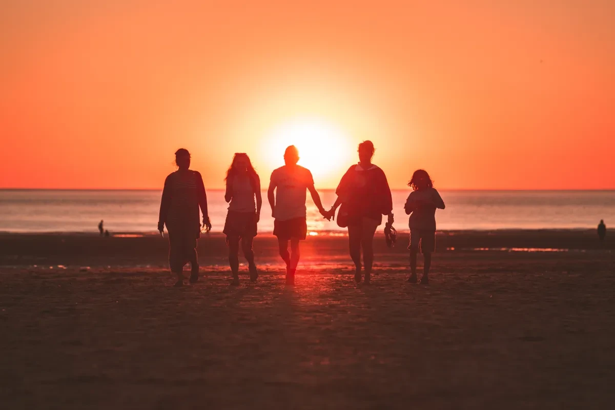 A family walks along a beach at sunset. The sun is setting behind them, framing them in silhouette