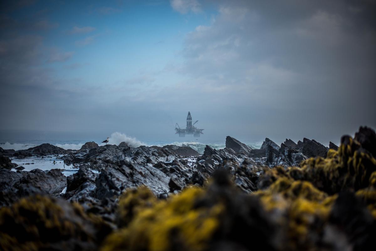 A photo taken from the shore of an offshore oil rig out to sea. The sky is grey and in the foreground waves are crashing against a rocky coast