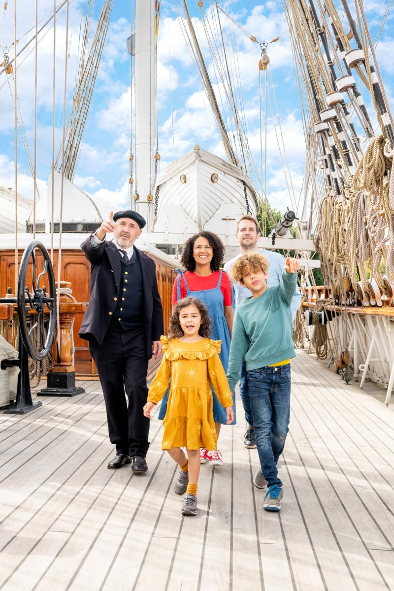 A family walks along the Main Deck of Cutty Sark, accompanied by a character actor dressed as the ship's captain. They are pointing to something behind the photographer, and the ship's rigging and lifeboat frames them in the background