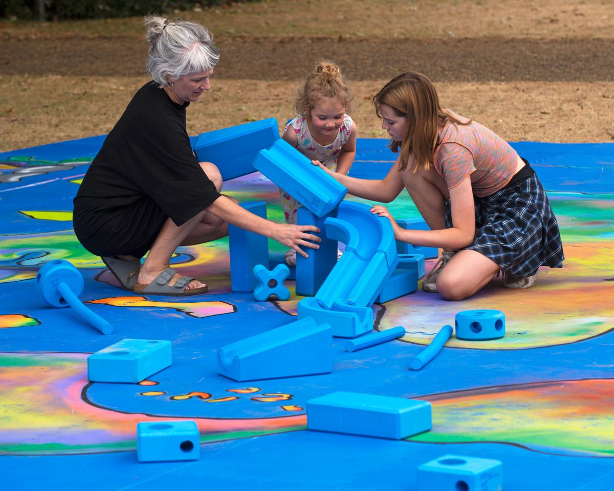 Two children and a young woman play with bright blue blocks as part of an outside play session at the National Maritime Museum