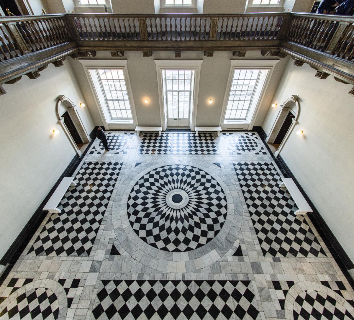 An overhead view of the Great Hall of the Queen's House, showing the geometric black and white pattern of the marble floor. Tall, ground level windows bathe the space in light, and a balcony runs round the top of the square room