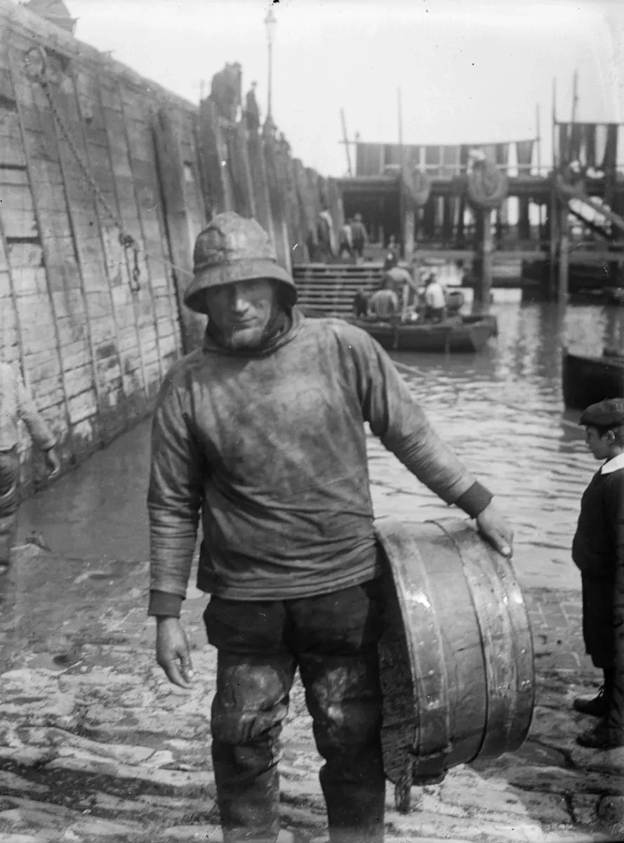 Historic black and white photograph of a fisherman in waders holding a barrel under his left arm. The harbour wall is visible behind his right shoulder