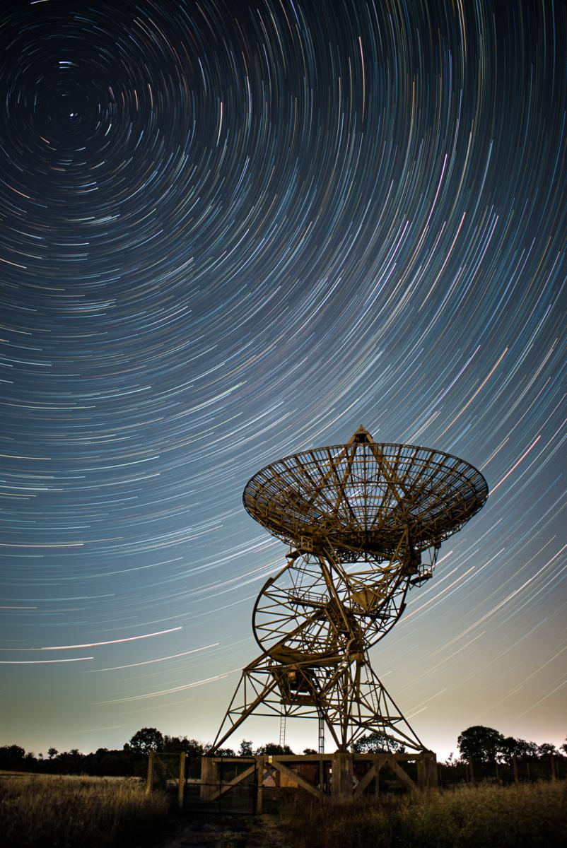 Image of radio telescope in the foreground and in the night sky behind it are semicircular star trails