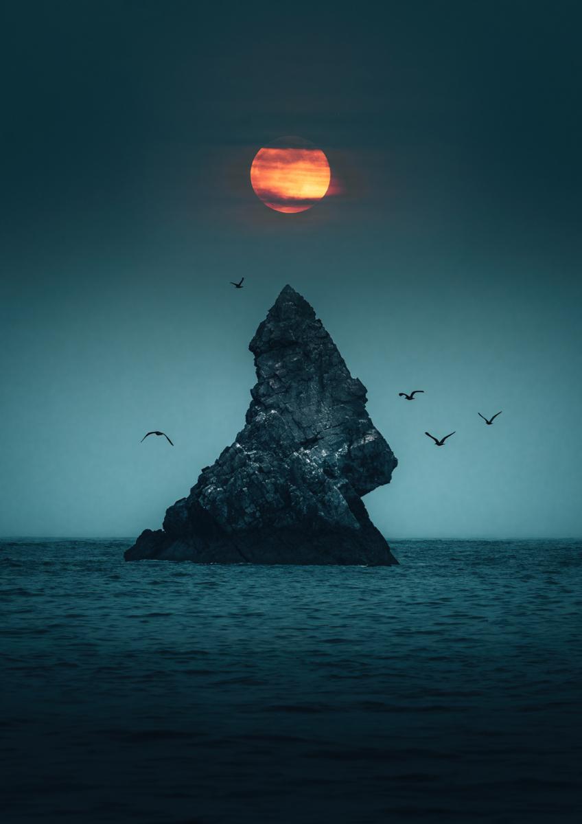 Image of a rocky formation in the sea, with the Moon behind it, slightly orange and obscured a bit by clouds