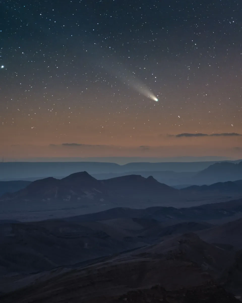 Image of Comet C/2021 A1 streaking through the sky leaving a long white tail, the sky is dusky as the sun sets