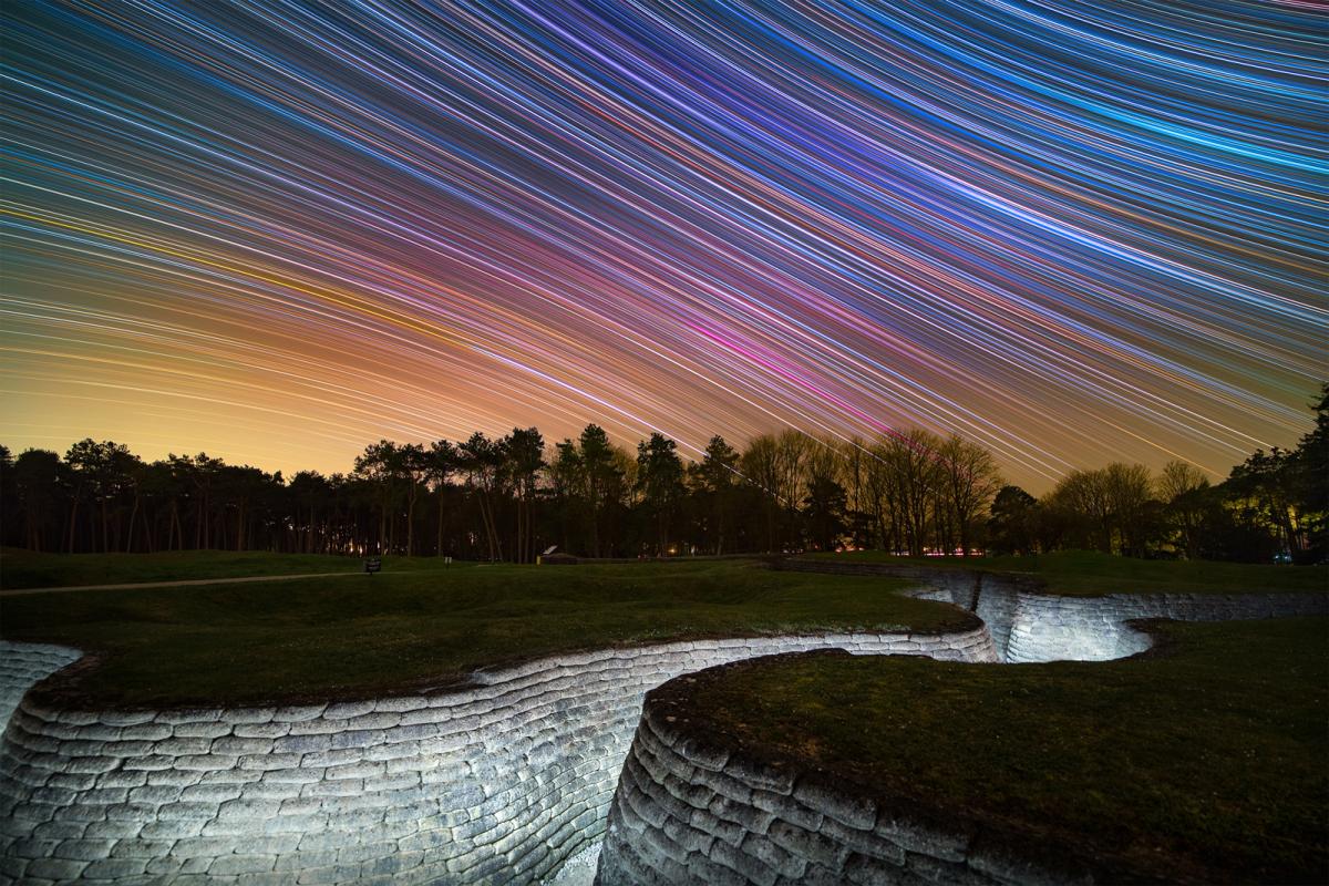 Image showing WWI trenches at bottom lit up with lights, a line of trees in the distance, and a purple, blue and yellow sky behind with star trails making curved diagonal lines