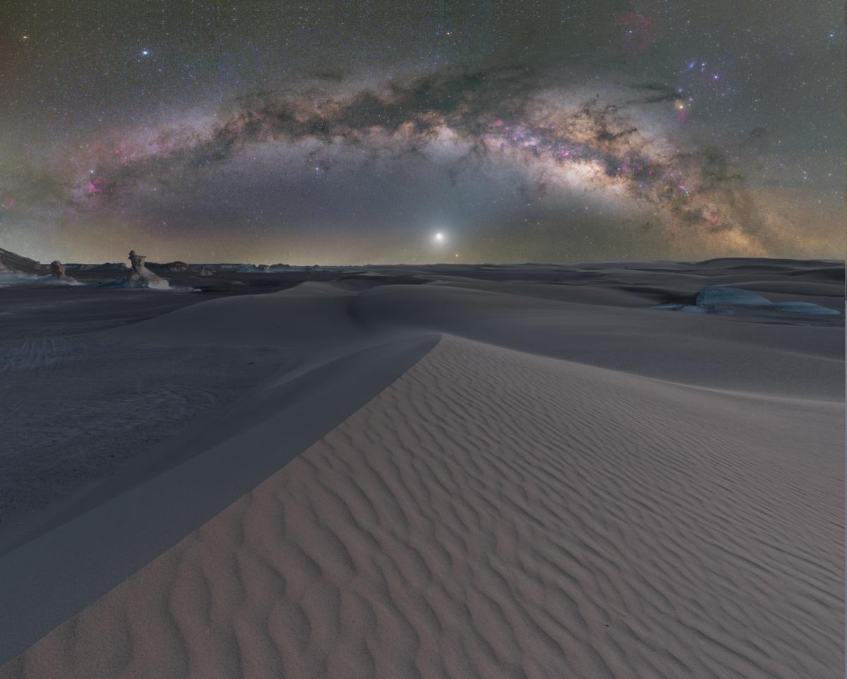 Image of sand dunes stretching into the horizon, above them Venus glows bright white in the middle of the image and the Milky Way curves over the top