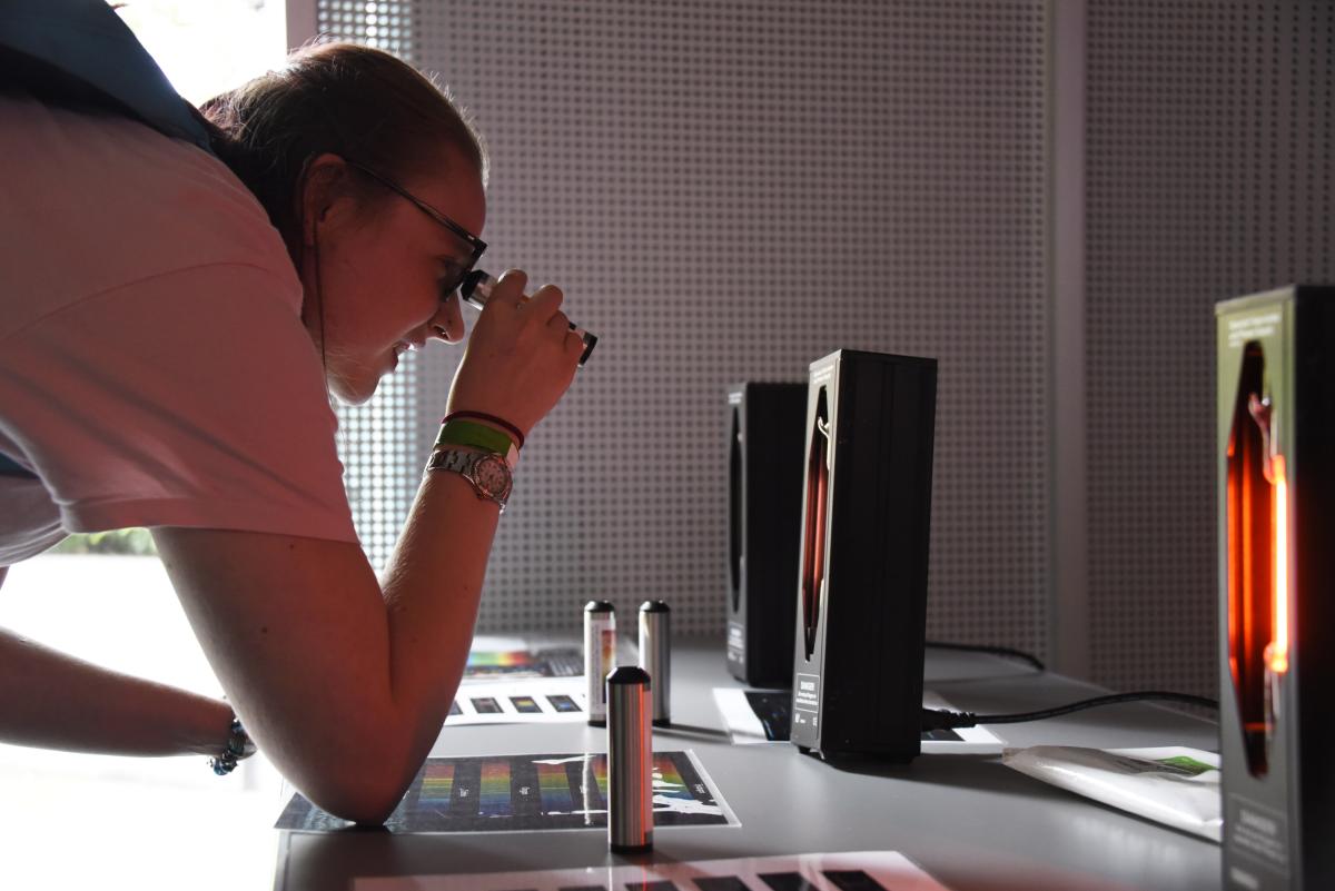 A woman looks through an eyepiece at a black box on a table emitting a red light. On the table in front of her are sheets of paper with the spectrum of light laid out in a bright rainbow of colour