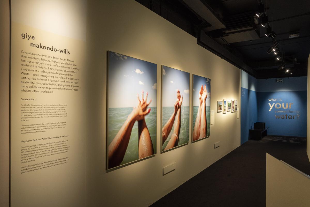 An interior gallery photograph of free exhibition Our Connection to Water at the National Maritime Museum. A display of photographs is on the wall