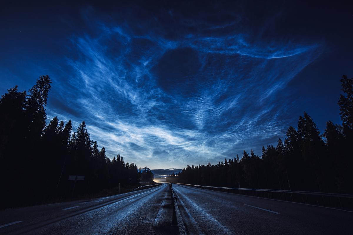 Image showing road at night with trees to the left and right, with sky above lit up with blue and silver noctilucent clouds