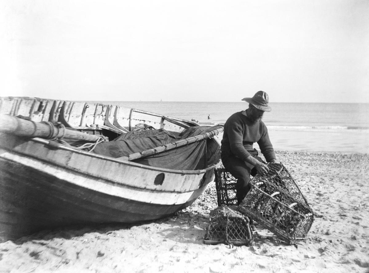 Black and white photograph showing A fisherman in sou'wester mending lobster/crab creels on the beach alongside a beached Sheringham crab boat 