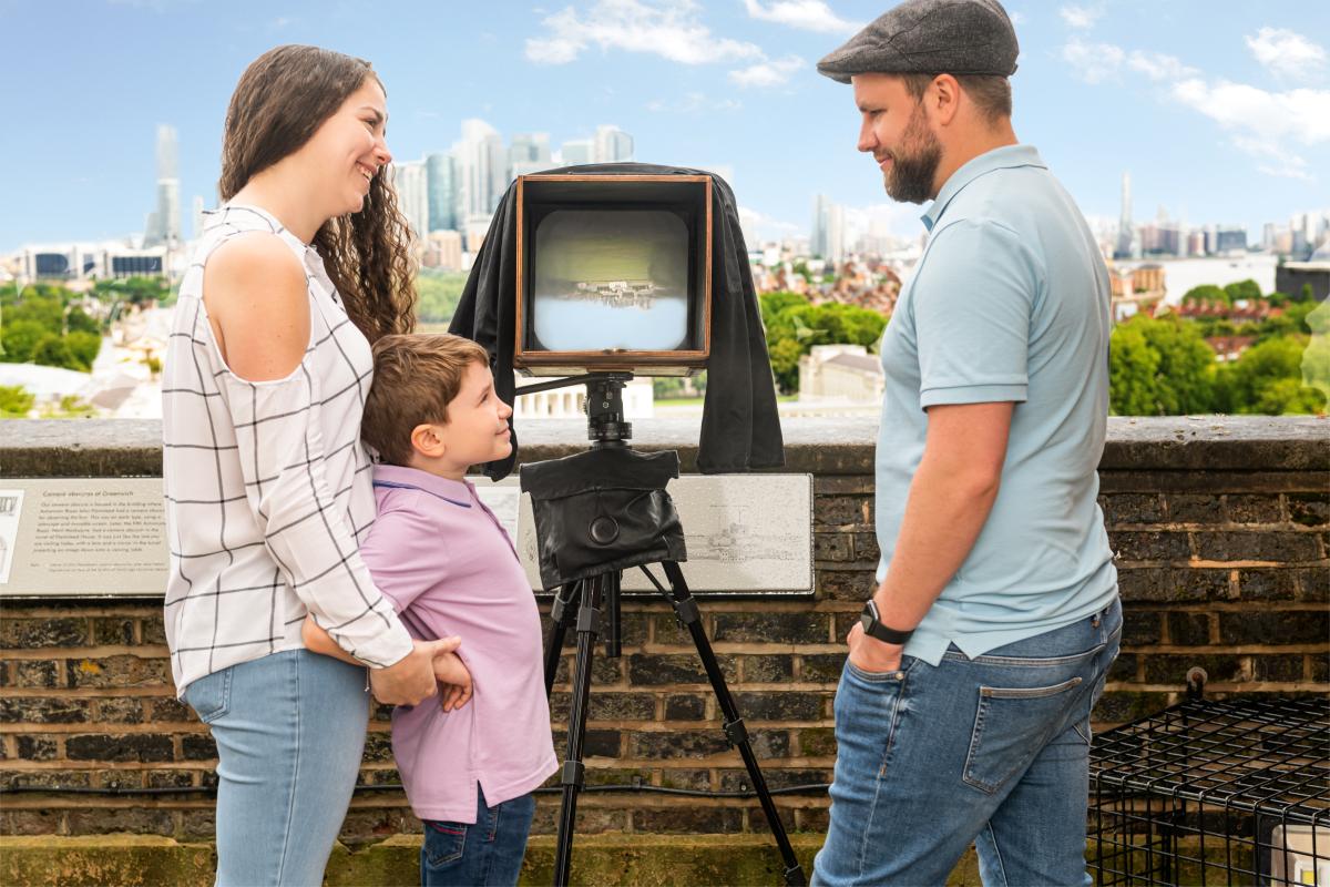 A woman, a man and a young boy look at a box and tripod containing a camera obscura at the Royal Observatory Greenwich. The viewing window of the camera shows an inverted image of the skyline behind them