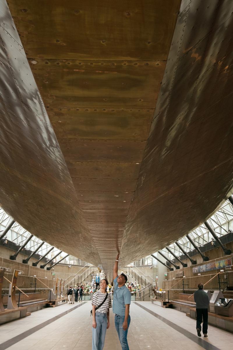 A couple look up at the hull of Cutty Sark. The ship appears to be floating above them, supported as it is by an array of steel beams