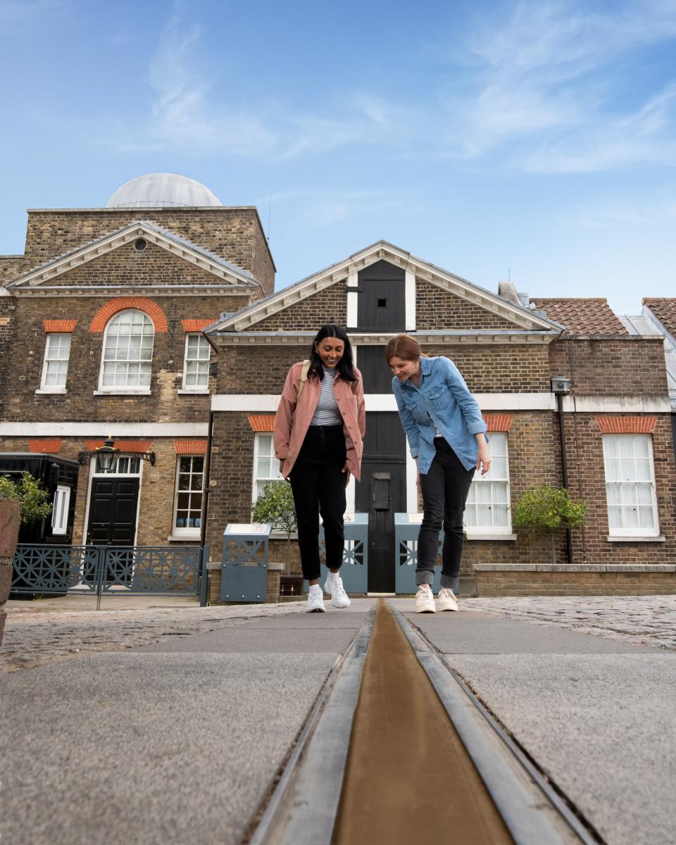 Two friends stand and point at the Prime Meridian Line in the courtyard of the Royal Observatory Greenwich
