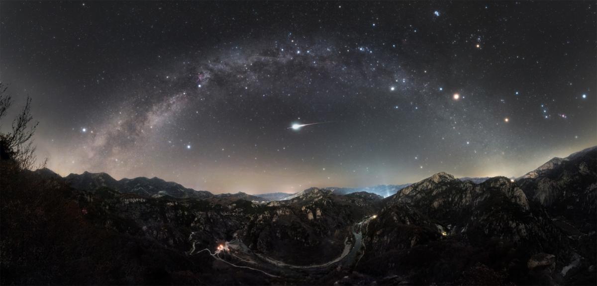 Rectangular image featuring a mountainous landscape in the bottom, with the Milky Way arching from the left to right of the image, smattered with stars. In the direct middle of the arc is an Orionid meteor, shining very brightly