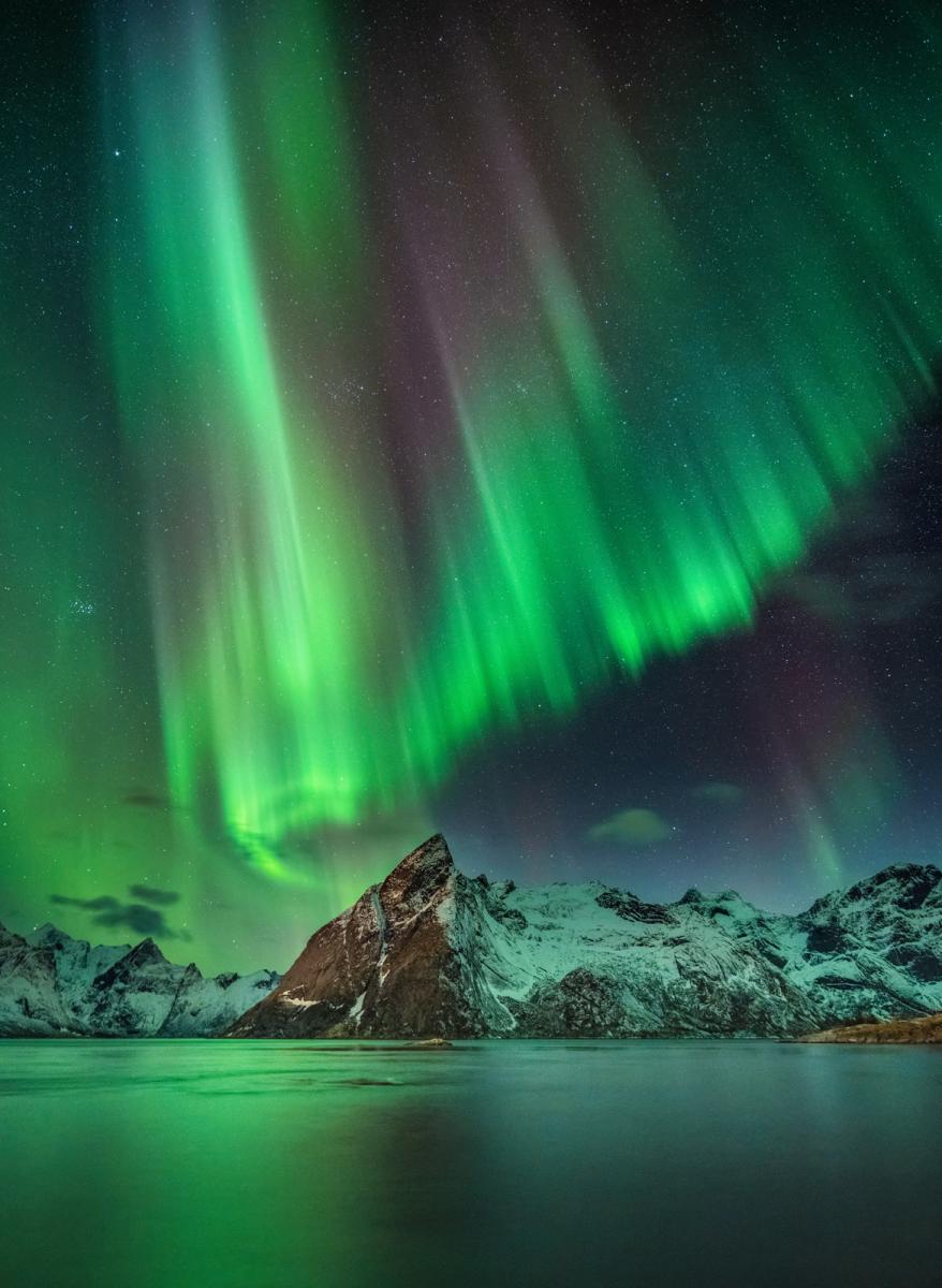 Image of snow-capped mountains next to a wide and still body of water, with aurorae in the sky in bright green hues that hang like a curtain over a starry night sky