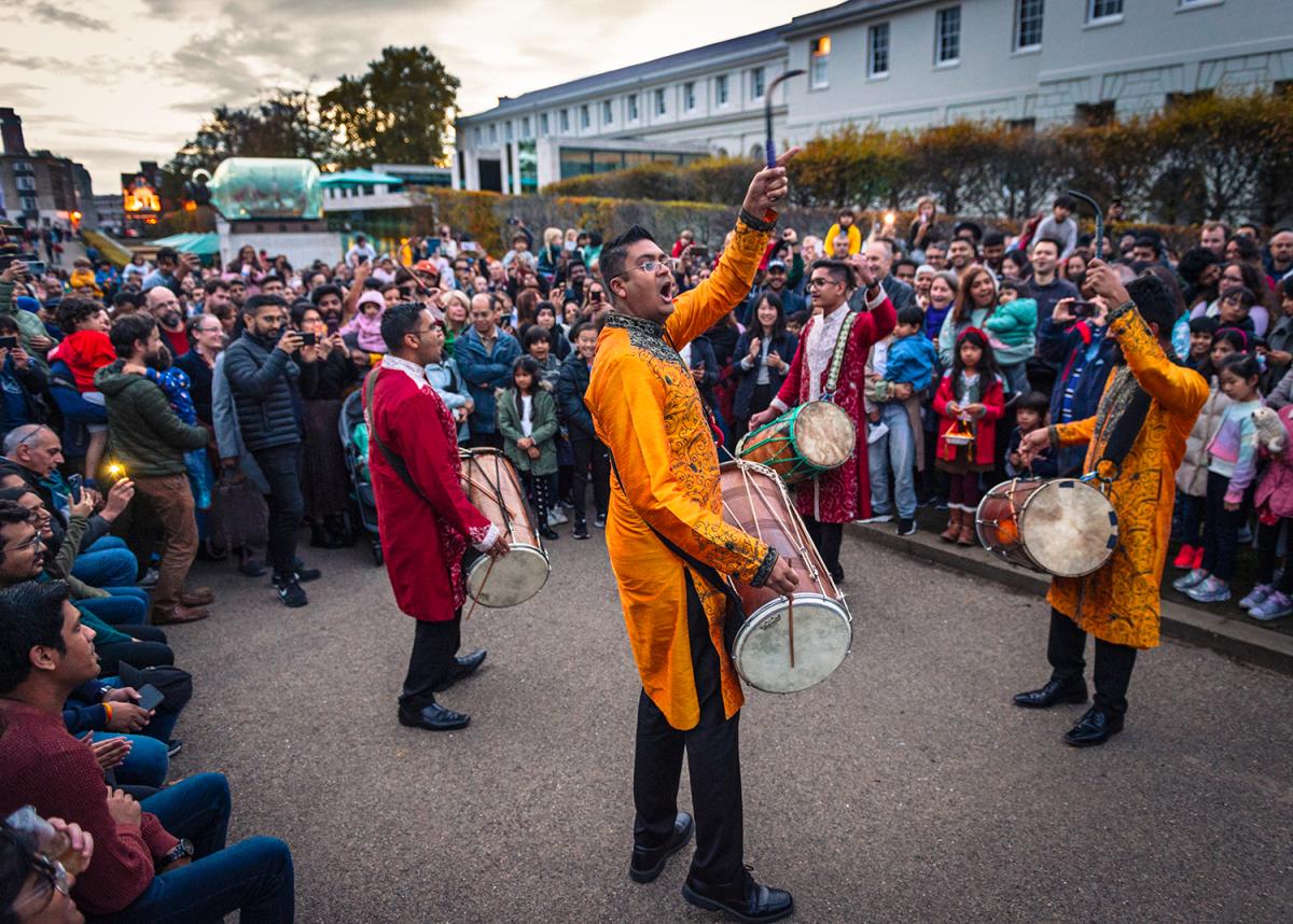 A group of south Asian drummers perform outside the National Maritime Museum in Greenwich at dusk, with a crowd looking on 