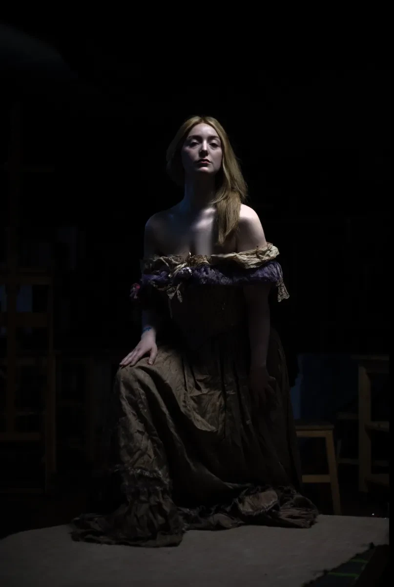 A photo of an actor playing Emma Hamilton. They kneel in the centre of a black background with long red hair falling over their shoulders. They wear a typical 19th century dress off the shoulders. 