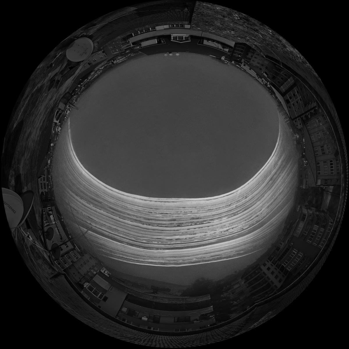 An artistic black and white fish eye view photograph, showing a cityscape as a 'circle' in the frame. In the middle of the circle, multiple bright white lines of light show where the Sun has passed on different days over the course of the photo's exposure