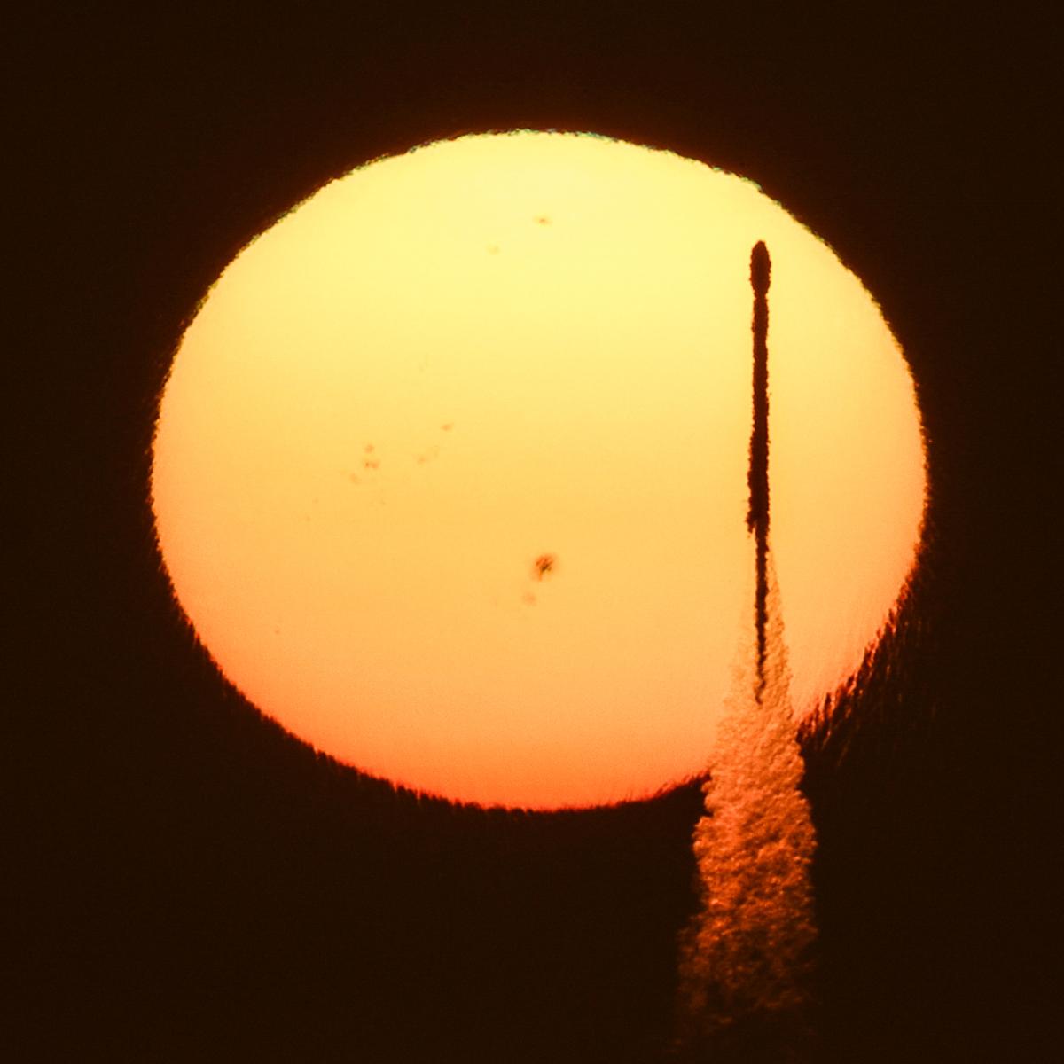 A dramatic photograph showing the launch of a SpaceX Falcon rocket. The rocket is picked out in dark silhouette against a setting Sun, with a hazy stream from the jet engines bursting out beneath like bubbles