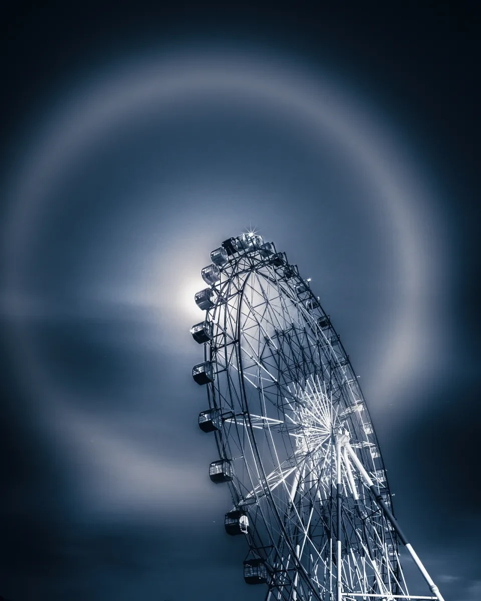 Image of a large ferris wheel from a ground angle, with the Moon just behind it, with a lunar halo which is a giant reflection ring around the Moon