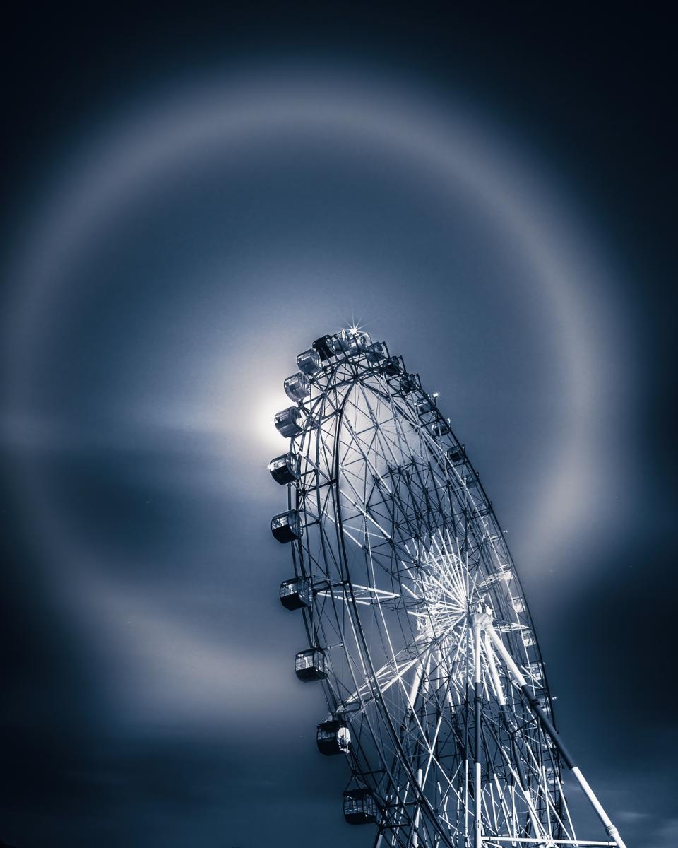 Image of a large ferris wheel from a ground angle, with the Moon just behind it, with a lunar halo which is a giant reflection ring around the Moon