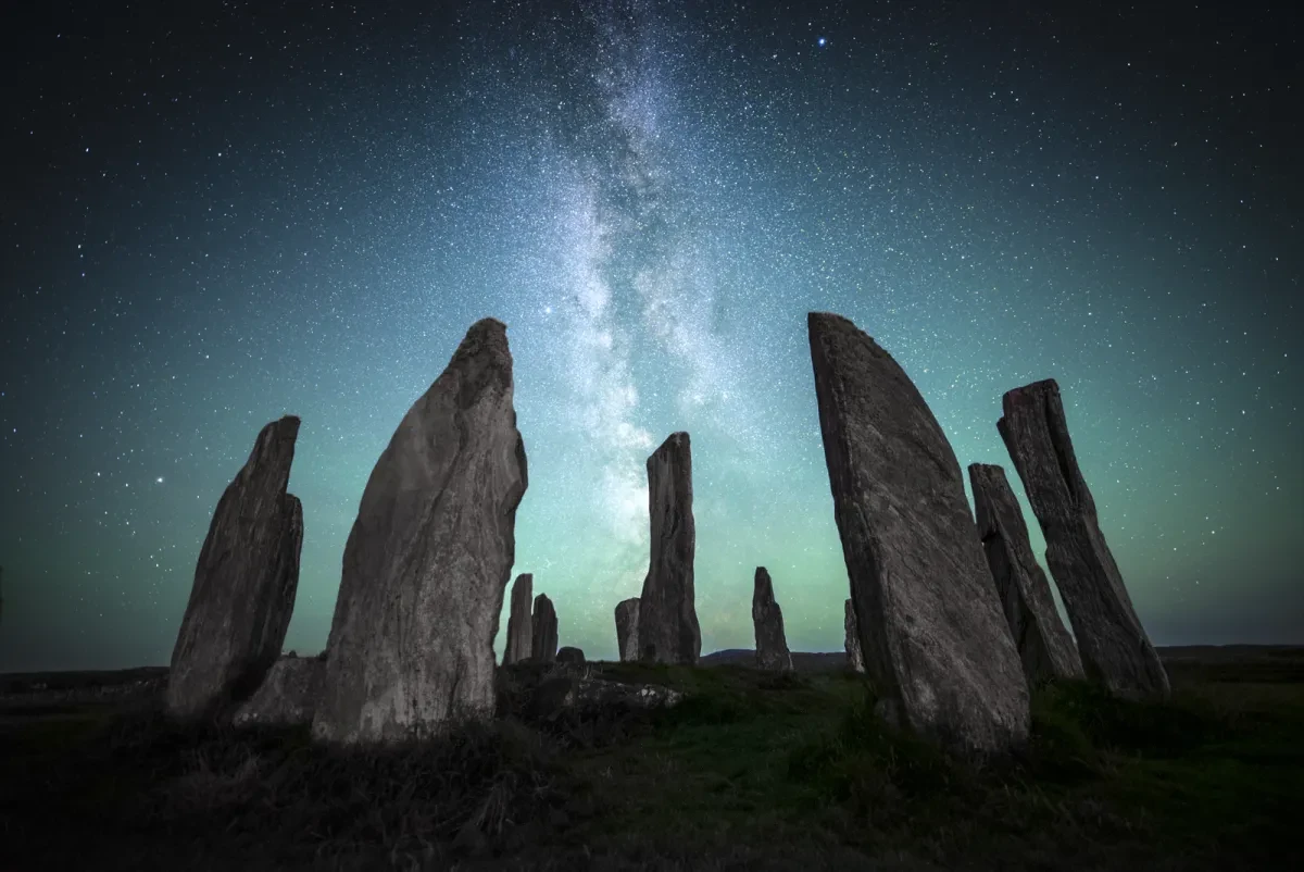 Image of a prehistoric rock formation akin to Stonehenge in the foreground of the image, with the Milky Way rendered in blues and greens in the sky
