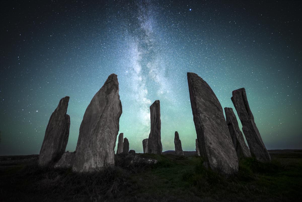 Image of a prehistoric rock formation akin to Stonehenge in the foreground of the image, with the Milky Way rendered in blues and greens in the sky