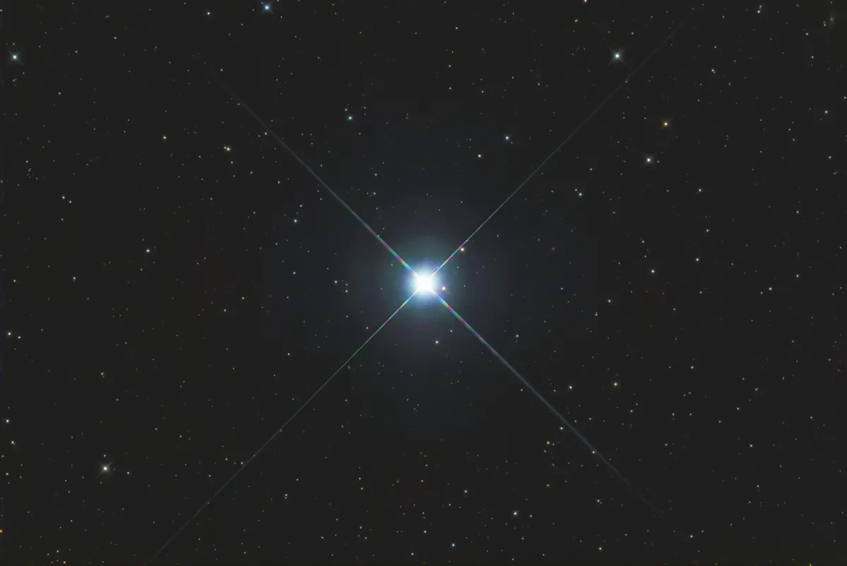 Image of a black starry night sky, with one very bright white and blue star in the centre with reflective lines coming off of it