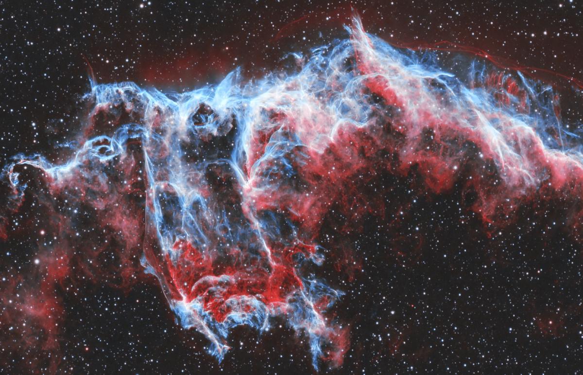 Image of a nebula resembling a wave in blues and reds mixed with an outline of white, against a black starry night sky