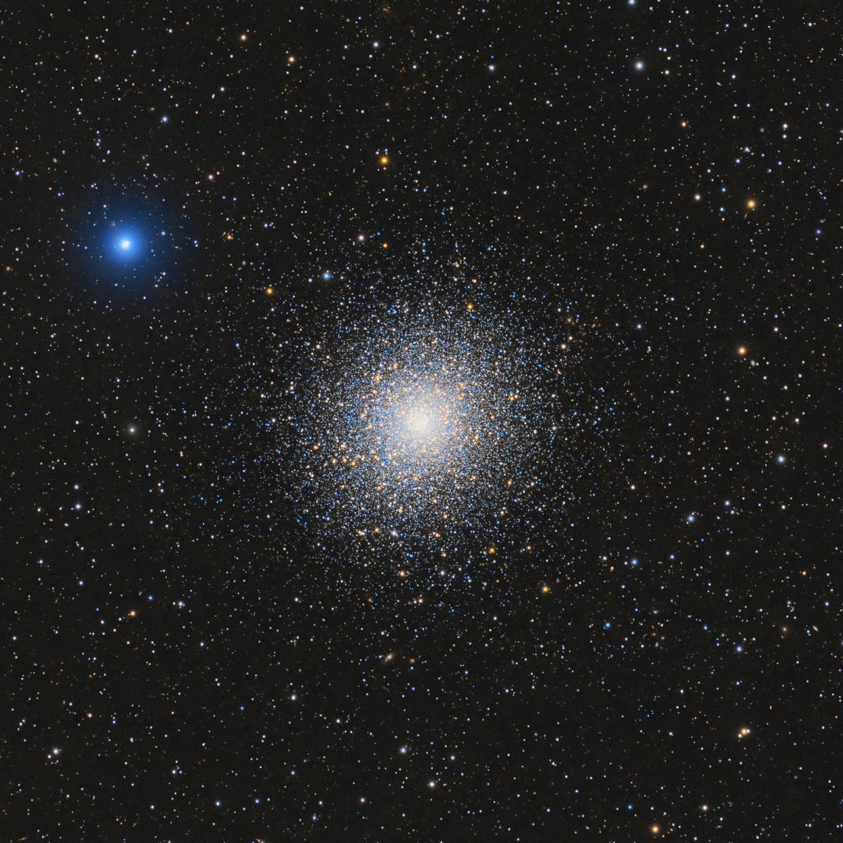 Against a black starry night sky filled with stars in blues, oranges and whites is a massive star cluster with thousands of stars all clumped together into a bright white centre