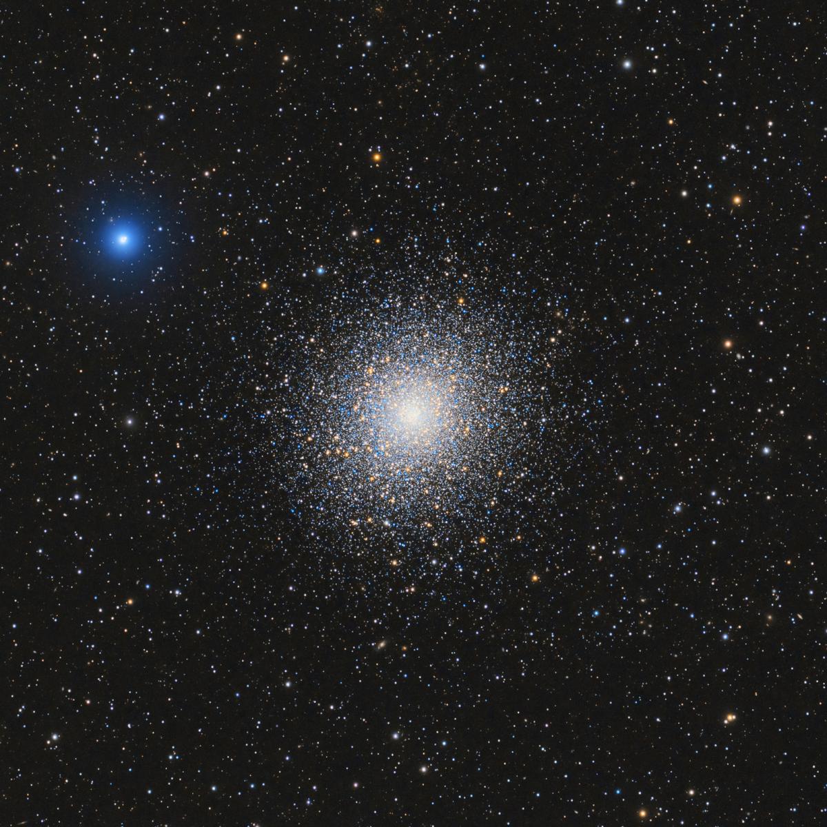 Against a black starry night sky filled with stars in blues, oranges and whites is a massive star cluster with thousands of stars all clumped together into a bright white centre