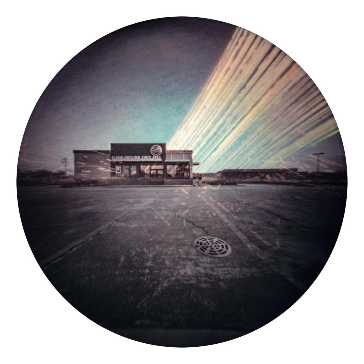 Circular solargraph image showing a fast food restaurant within a parking lot and in the sky is multiple pale yellow lines moving from behind the restaurant to the upper right hand corner