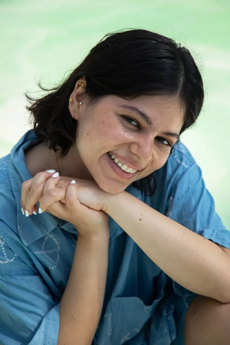 A smiling woman with her head to one side, resting her chin against her clasped hands. She has straight black hair, brown skin and is wearing a loose blue shirt