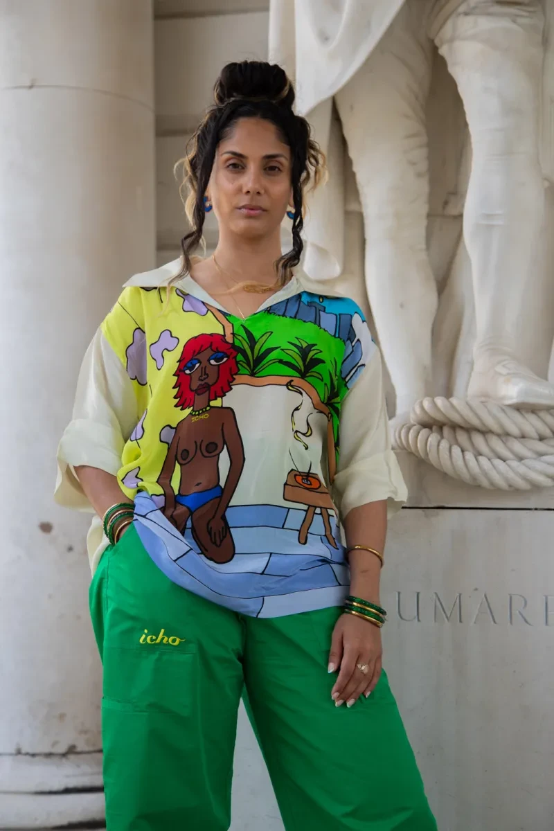 Portrait photo of a woman in brightly patterned clothing. Her long dark hair is partially up in a bun and she is staring confidently down at the camera. Her loose shirt features a cartoon-style artwork of a naked woman kneeling next to an open window