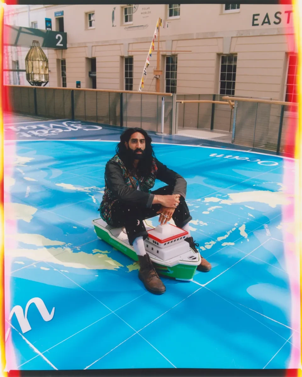 Photograph of a man sitting on a miniature ride-on ship in a Museum. The floor is styled to look like a giant map. The man has a dark, well-trimmed beard, long black hair and brown skin. He looks thoughtfully up at the camera