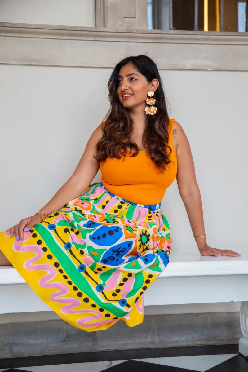 Portrait photograph of a woman with long brown hair, wearing a bright orange top and multicoloured patterned dress. She is looking to her right, and her hair is tucked behind her left ear. In the ear she is wearing a striking hanging earring