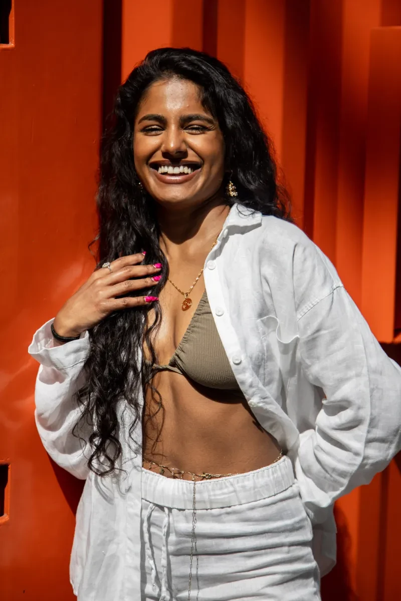 Photograph of a laughing woman with long dark hair and brown skin. She is wearing white trousers and a loose fitting, unbuttoned white shirt, with a dark green bikini top underneath