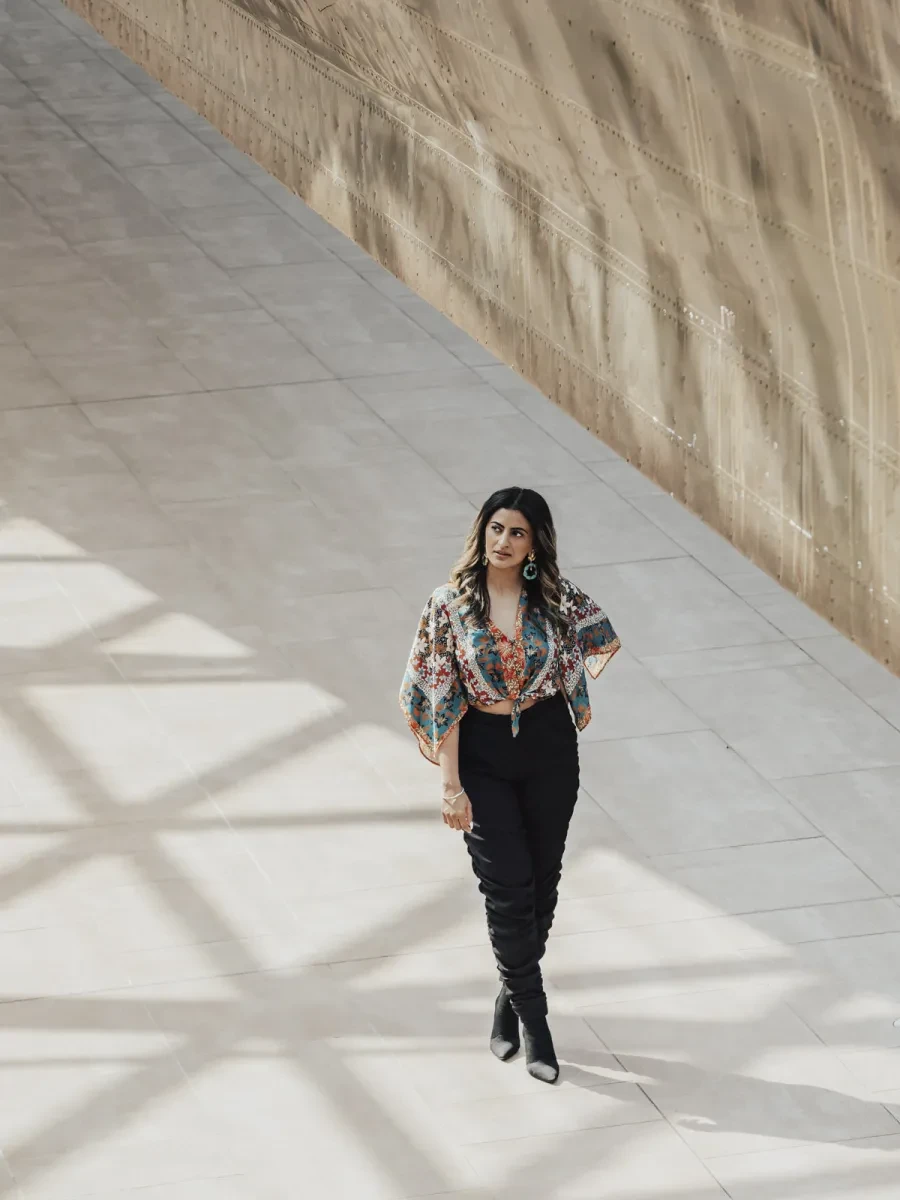 Photograph of a woman walking in a brightly lit, stone-floored space. She has long brown hair, dark jeans and a brightly patterned fabric top