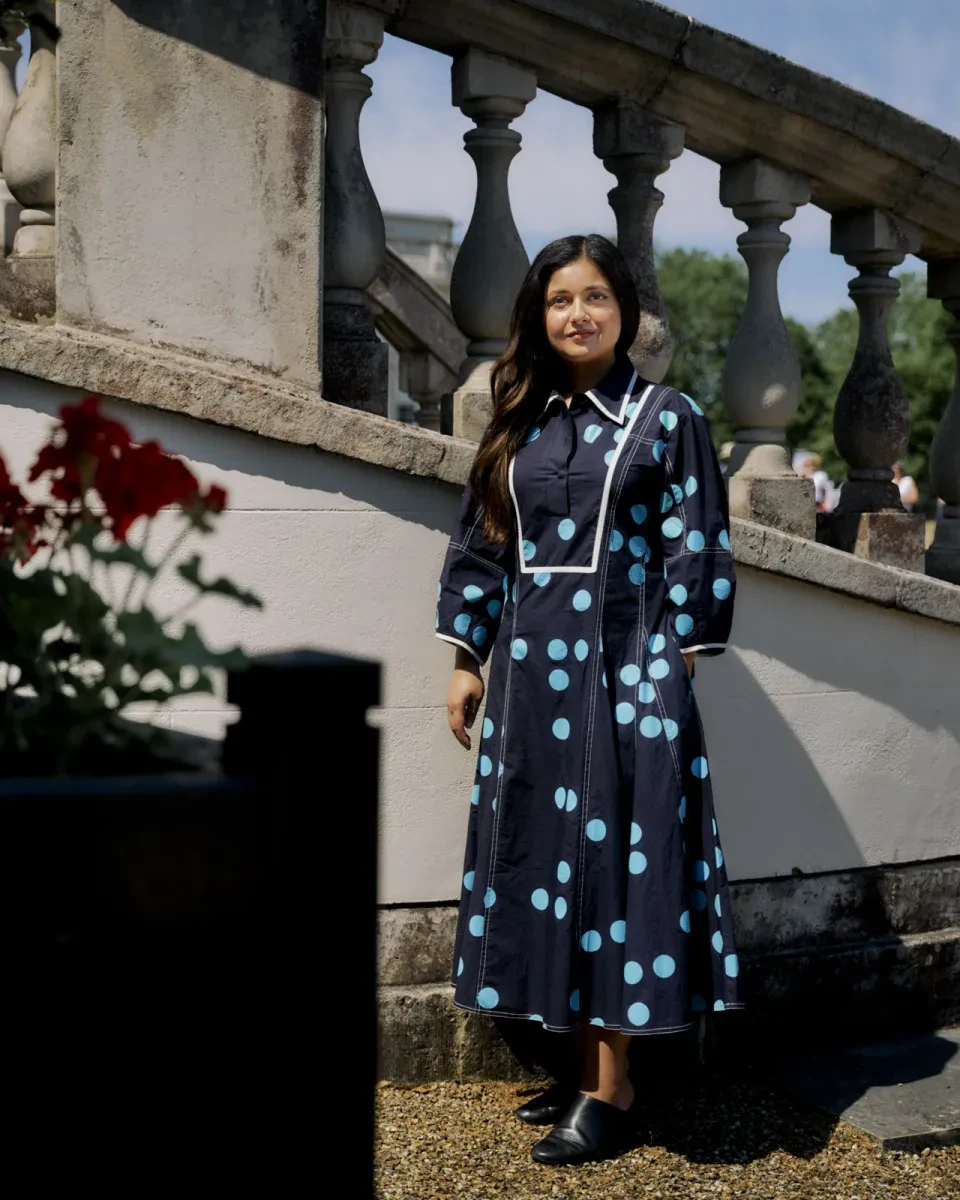 Full length photograph of a woman wearing a striking dark blue dress with bright blue polkadots. Her hair is long, dark and wavy, and hangs neatly over her right shoulder