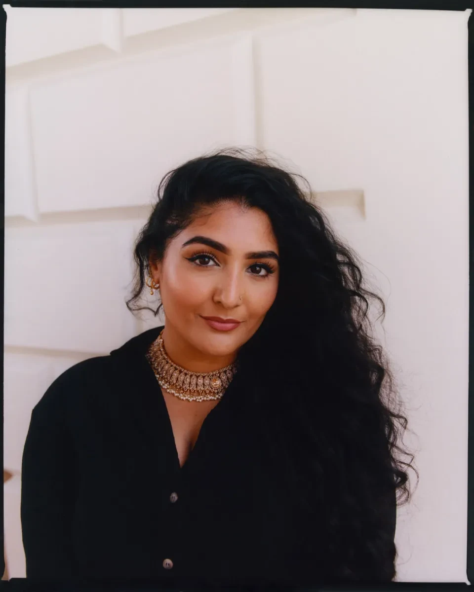 Portrait photograph of a young woman with light brown skin and long black curly hair. She is looking confidently at the camera, with one eyebrow slightly raised