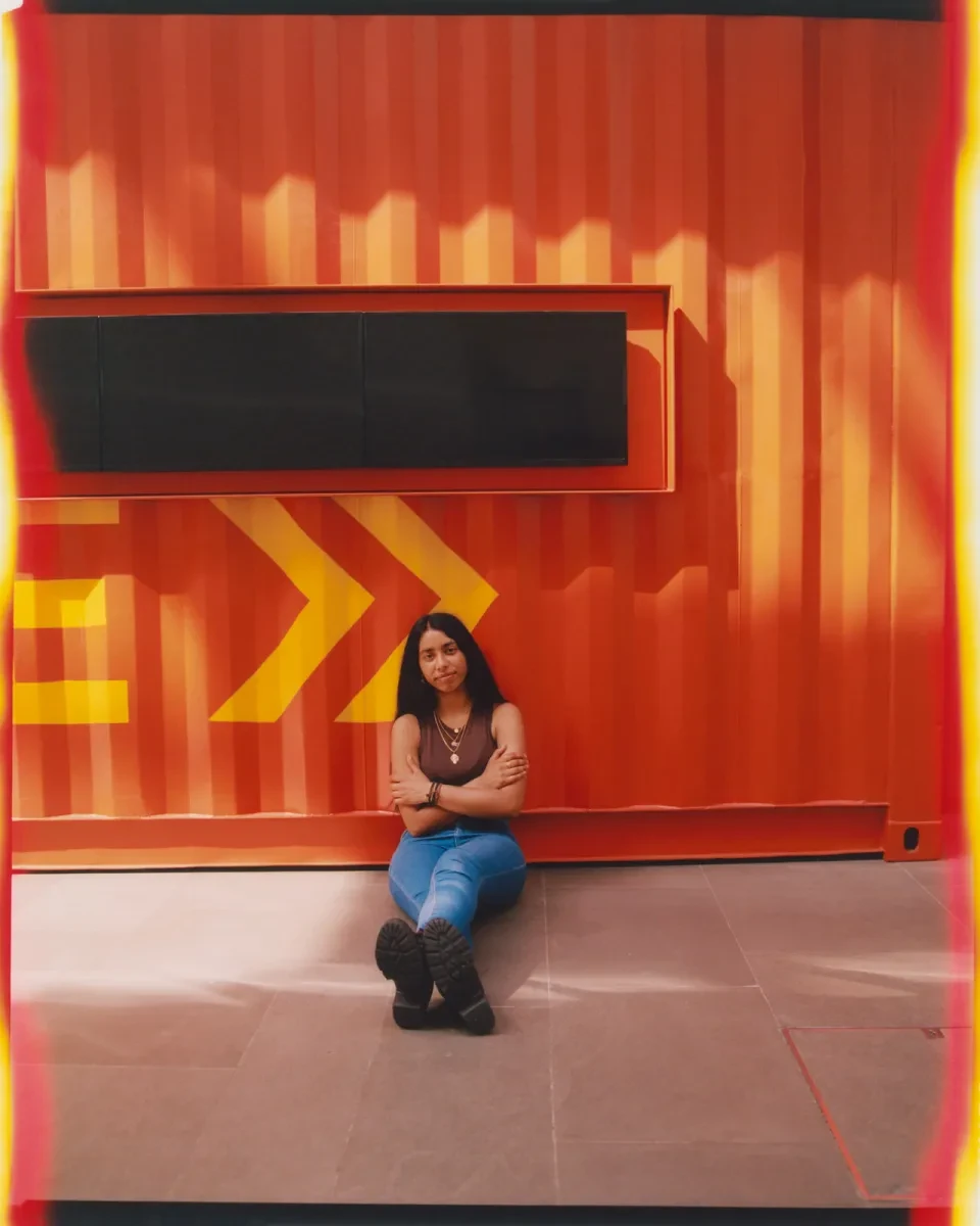 Photograph of a woman sitting on the ground next to an orange shipping container. She has her arms folded and is leaning against the corrugated metal