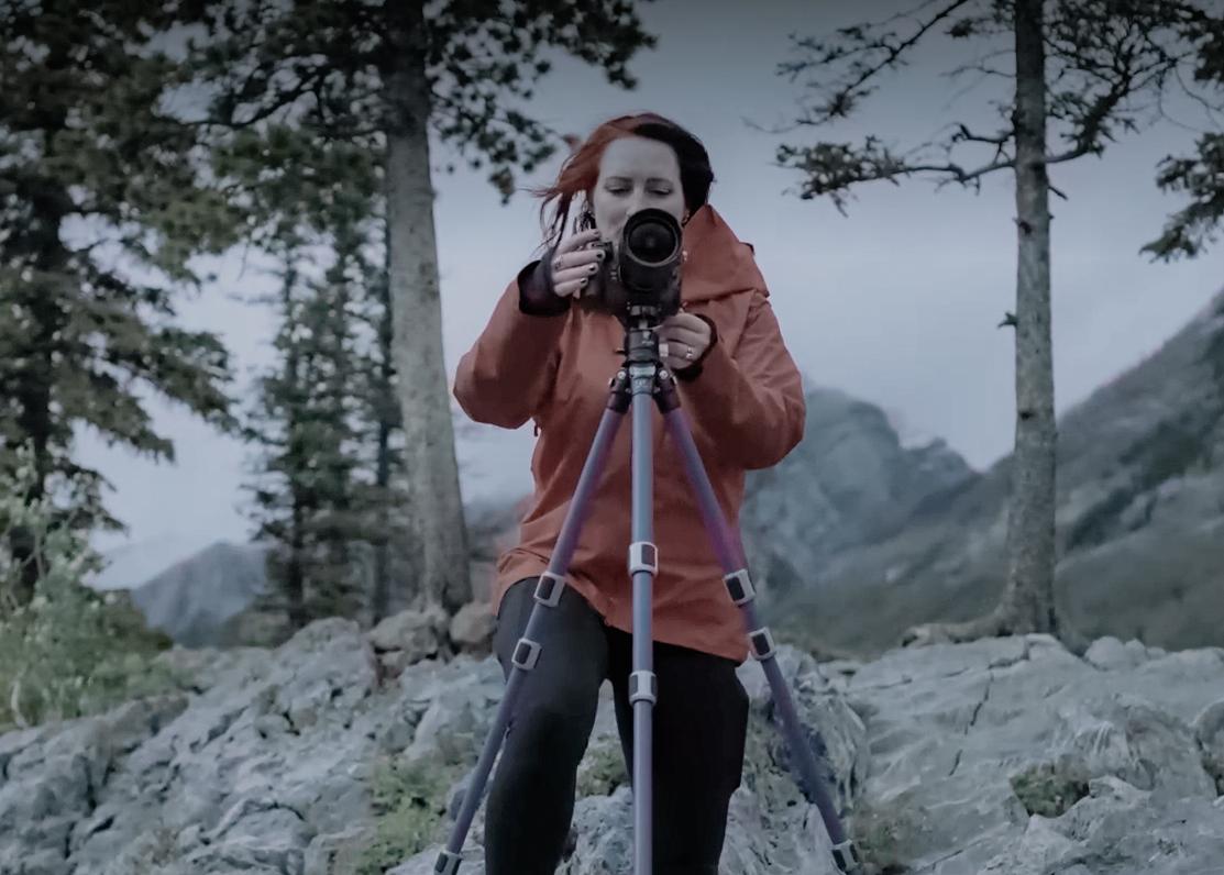 A woman takes a photograph using a DSLR and a tripod. She is standing in a forest in front of grey rocks, wearing an orange-red coat, her camera facing the viewer
