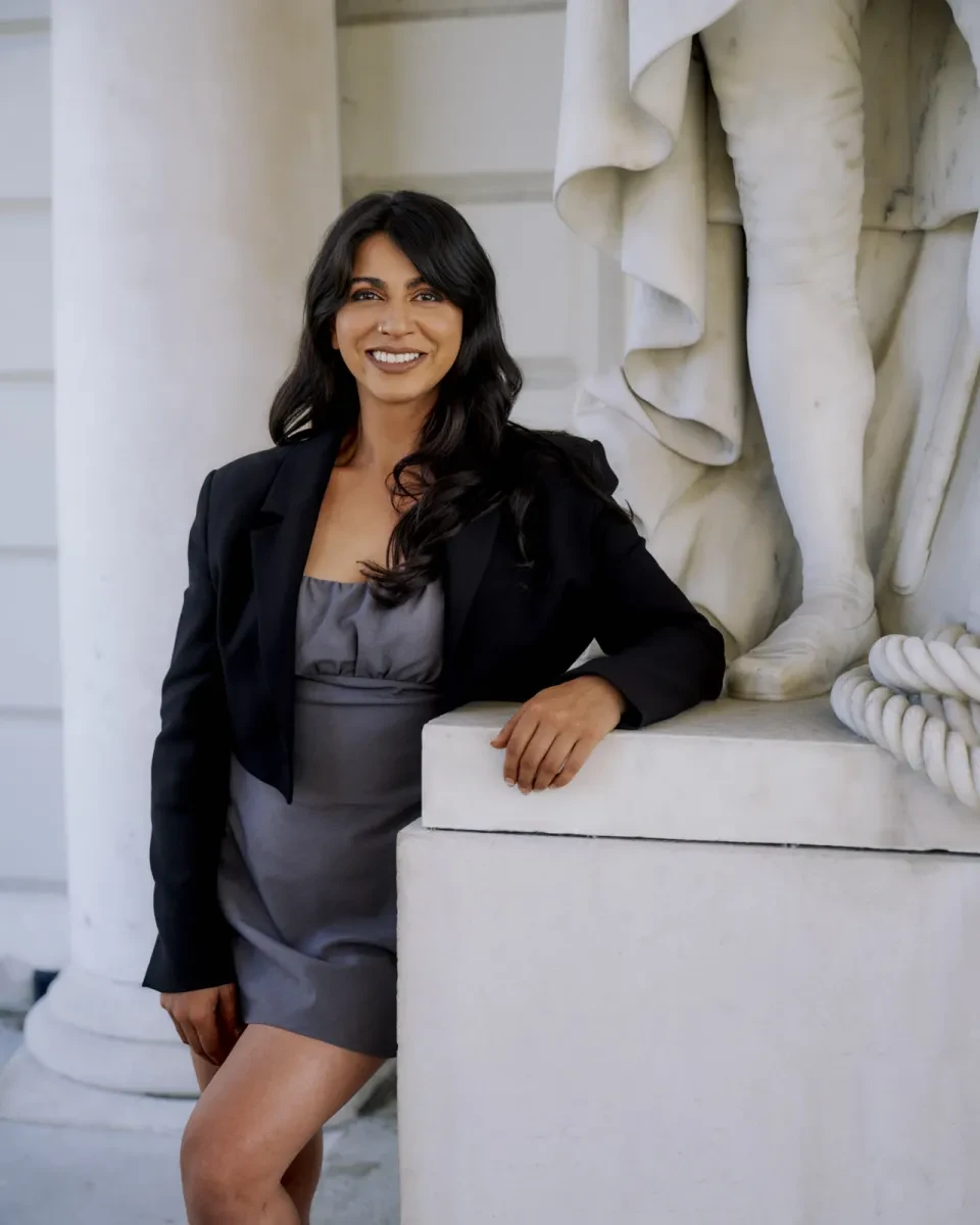 Portrait photograph of a woman wearing a black blazer and short grey dress. She has long wavy black hair and is smiling brightly directly at the camera