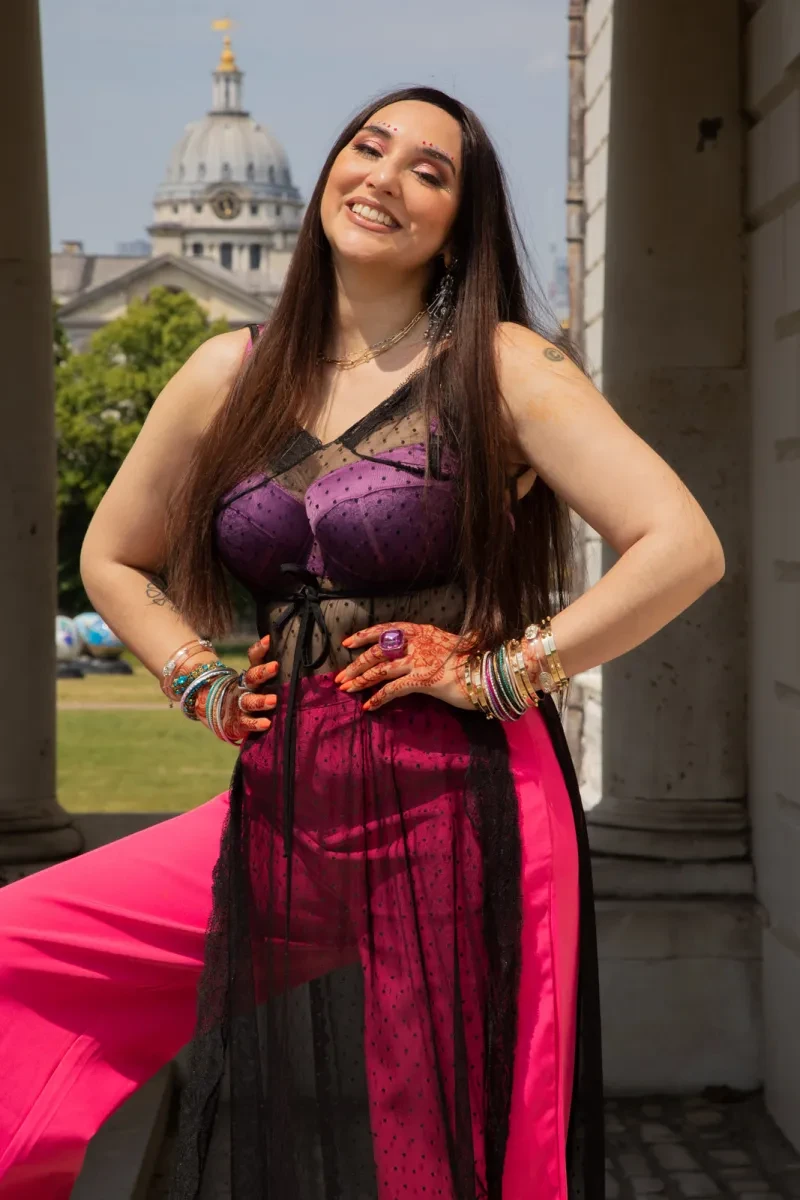 Portrait photograph of a woman smiling outside historic building in Greenwich. She is wearing pink trousers and a sheer black top with a purple bra visible beneath. Her hands are on her hips, and her fingers are covered in intricate henna designs