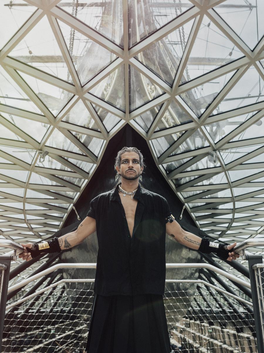 Striking portrait photograph of a man standing beneath historic ship Cutty Sark. He is wearing a black loose shirt with fingerless gloves, wears heavy eyeliner and a thick silver necklace.