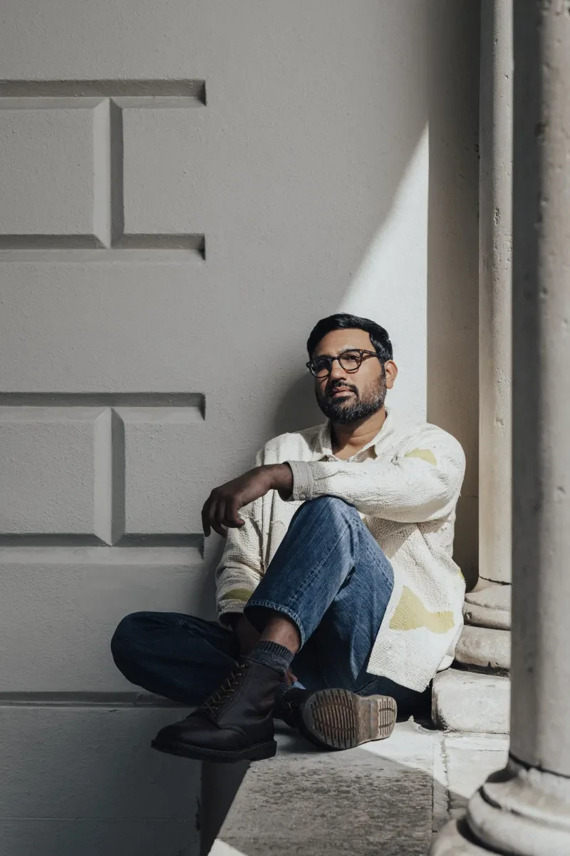 A man sits contemplatively next to a grey stone wall. He has a dark stubbly beard and is wearing glasses. A shaft of sunlight illuminates his face and left shoulder